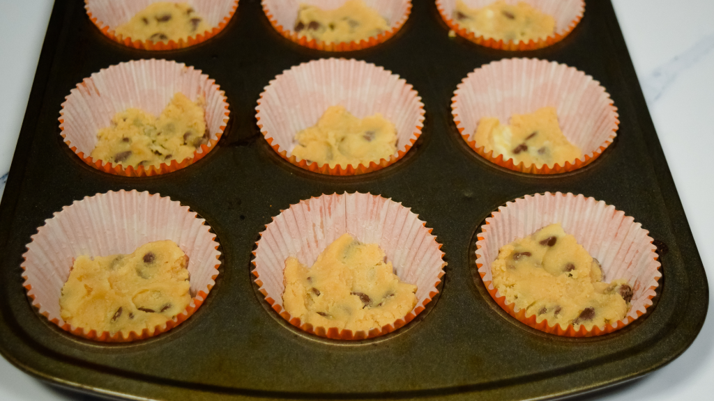 A muffin tin with orange cupcake liners and each liner has chocolate chip cookie dough in it