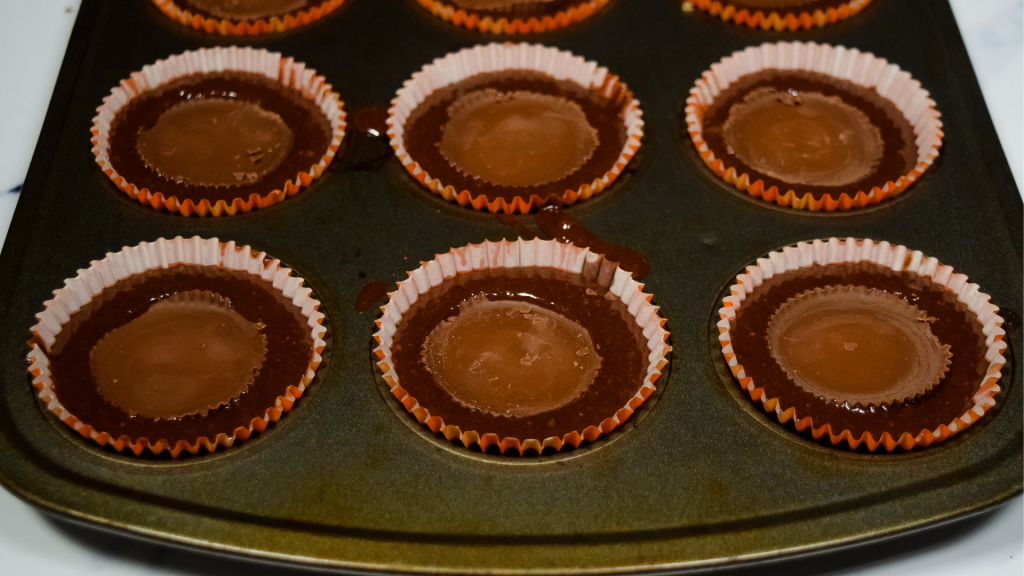 A muffin tin with orange cupcake liners with peanut butter cups sticking out of brownie batter. 