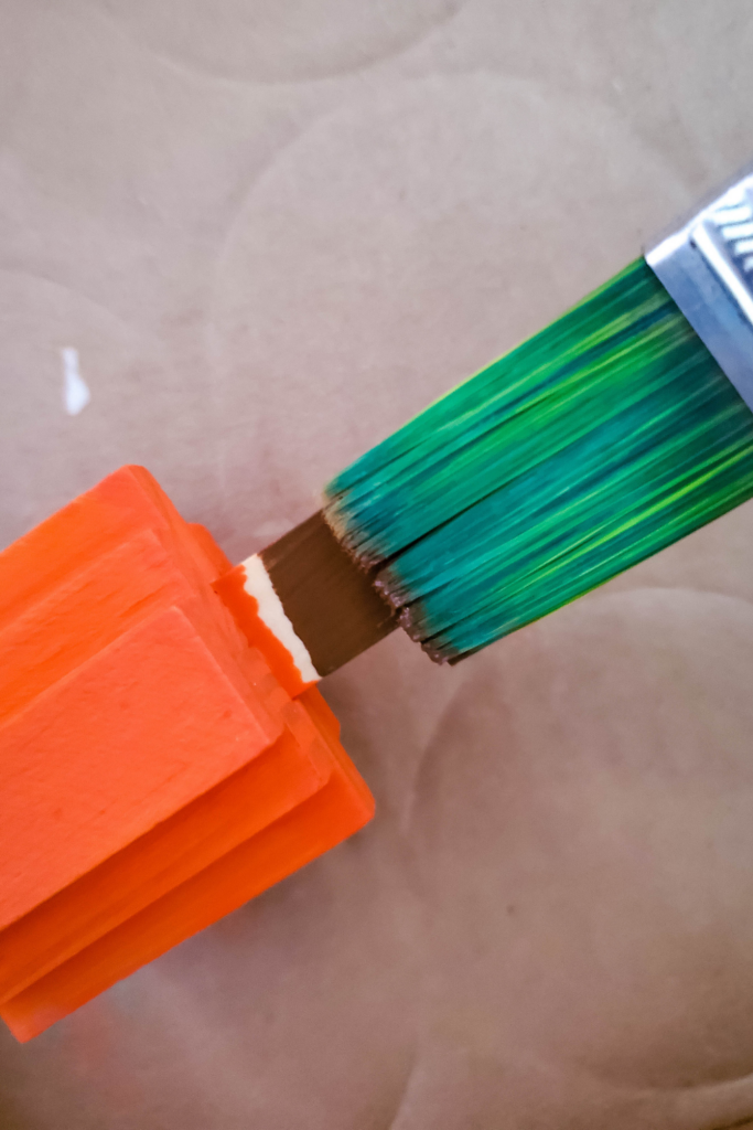 A green paint brush painting the stem of the jenga block pumpkin brown. 